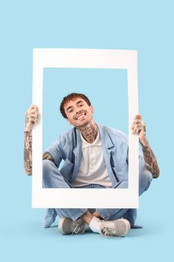 Mavi arka planda beyaz fotoğraf çerçevesi tutan mutlu genç adam.