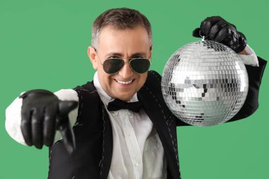 Havalı, takım elbiseli, disko toplu olgun bir adam yeşil arka plandaki izleyiciyi işaret ediyor.