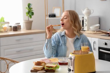 Genç ve güzel bir kadın reçelli tost yiyor ve mutfakta portakal suyu içiyor.