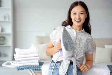 Evde ütü masasının yanında demirli mutlu bir Asyalı kadın var.