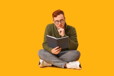 Genç adam sarı arka planda oturuyor ve kitap okuyor.