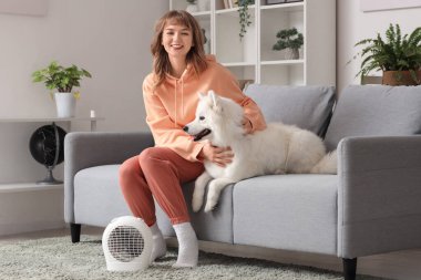 Samoyed köpeğiyle evdeki elektrikli vantilatör ısıtıcısının yanında ısınan genç kadın.