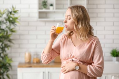 Genç hamile kadın mutfakta portakal suyu içiyor.
