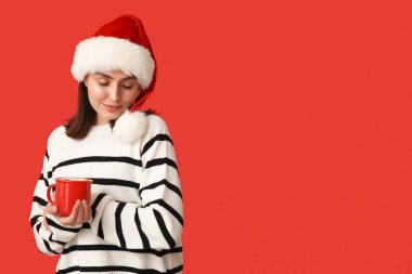Kırmızı arka planda sıcak şarap bardağıyla Noel Baba şapkalı genç bir kadın.