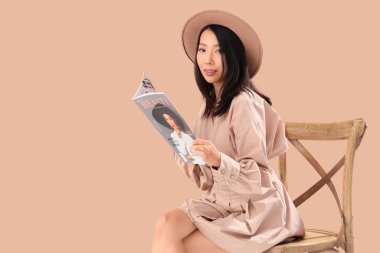 Genç Asyalı kadın bej arka planda sandalyede dergi okuyor.