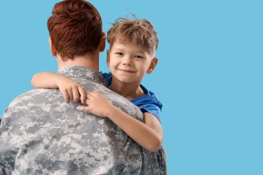 Küçük çocuk mavi arka planda askeri babasına sarılıyor.