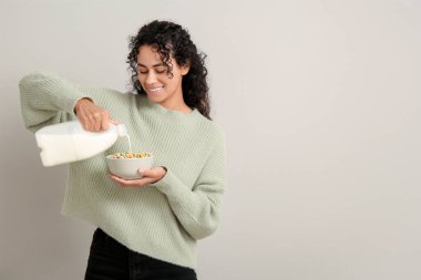 Smiling young woman pouring milk into bowl of cereal rings on light background