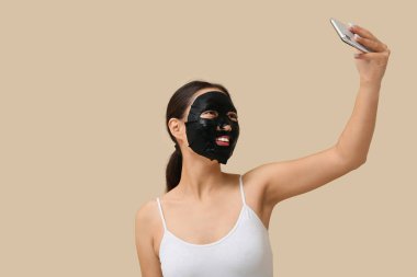 Yüzünde aktif kömür maskesi olan genç bir kadın bej arka planda selfie çekiyor.