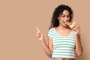 Güzel, genç, mutlu, Afro-Amerikan bir kadın. Bir dilim lezzetli pizzayla bej arka plandaki bir şeyi işaret ediyor.