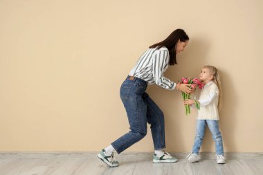 Küçük kız, mutlu annesini bej duvarın yanında bir buket çiçekle tebrik ediyor. Anneler Günü kutlaması