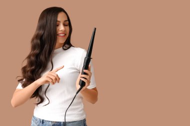 Beautiful young woman with curling iron on beige background