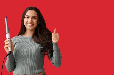 Kırmızı arka planda baş parmağını gösteren saç maşası olan güzel genç bir kadın.