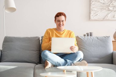 Genç adam oturma odasındaki koltukta dizüstü bilgisayar kullanıyor.