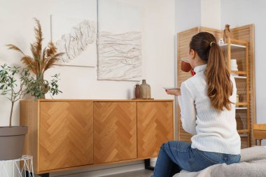 Elinde bir fincan kahveyle yatak odasındaki 3 boyutlu resimlere bakan genç bir kadın.