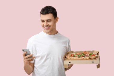 Elinde pembe arka planda lezzetli pizza ve cep telefonu olan karton bir kutu tutan genç adam.