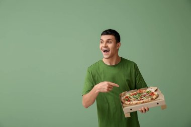 Genç adam yeşil zemin üzerinde lezzetli pizzalar olan karton kutuyu işaret ediyor.
