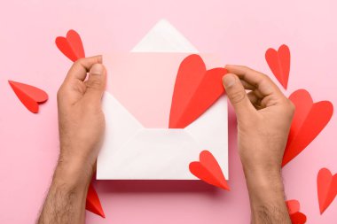 Zarflı erkek eller, boş kart ve pembe arka planda kırmızı kağıt kalpler. Sevgililer Günü kutlaması