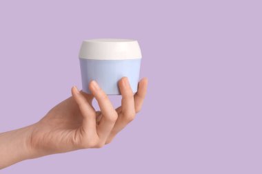 Leylak arka planında doğal krema kavanozu olan kadın eli, yakın plan.