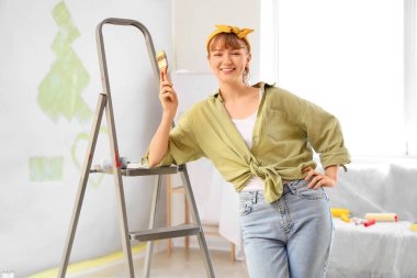 Boya fırçası ve merdiveni olan genç bir kadın odada tamirat yapıyor.