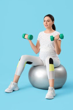 Güzel, genç, hamile bir kadın, spor yapıyor ve mavi arka planda dambıllar var.