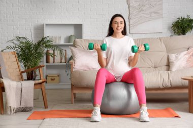 Güzel, hamile bir kadın evde spor salonu ve dambıllarla antrenman yapıyor.