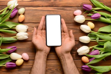 Modern cep telefonlu erkek eller, güzel lale çiçekleri ve tahta arka planda Paskalya yumurtaları.