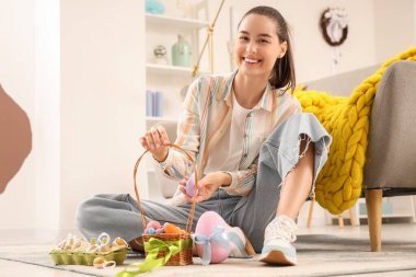 Paskalya yumurtası dolu sepeti olan genç bir kadın evde yerde oturuyor.