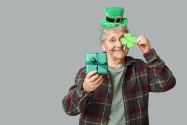 Cüce şapkalı, yonca ve gri arka planda hediye kutusu olan yaşlı bir kadın. Aziz Patrick Günü kutlaması