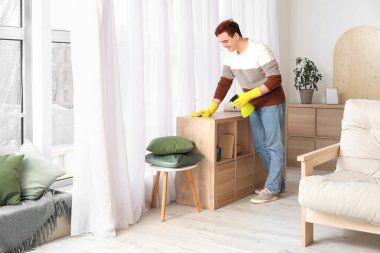 Oturma odasında paçavra ve sprey boyayla temizlik yapan genç bir adam.