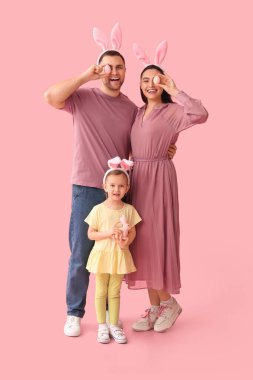 Tavşan kulaklı mutlu bir aile pembe arka planda Paskalya yumurtası tutuyor.