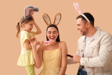 Paskalya tavşanı kulaklarında babasıyla küçük bir kız bej arka planda annesini gıdıklıyor.