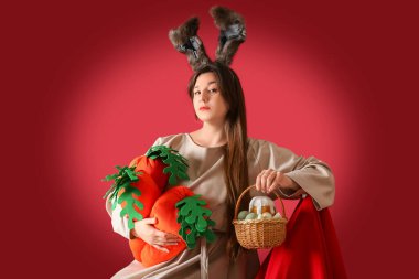 Paskalya tavşanı kulaklı genç bir kadın kırmızı arka planda paskalya yumurtası ve peluş havuç ve sepeti tutuyor.