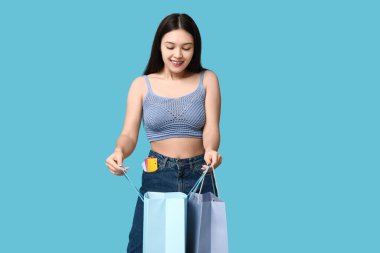Kot etekli, şık bir Asyalı kadın alışveriş torbaları ve mavi arka planda kredi kartları var.