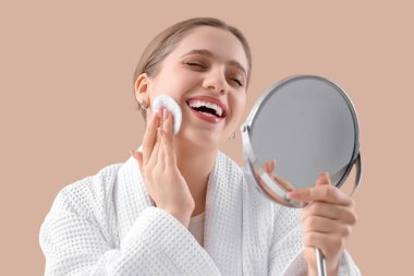 Bornozlu, pamuklu ve aynalı güzel bir kadın. Bej arka plandaki makyajı kaldırıyor. Yakın plan.