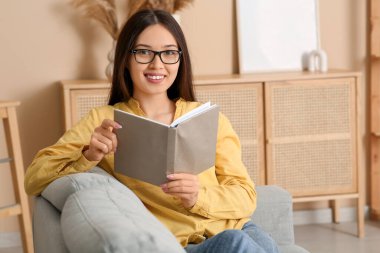 Güzel, genç Asyalı, gözlüklü, kitaplı bir kadın evdeki kanepede oturuyor.