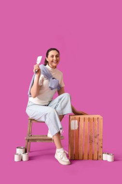 Genç bir kadın mor arka planda ahşap kutu boyuyor.