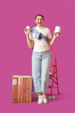 Teneke kutuları olan genç bir kadın mor arka planda tahta kutu boyuyor.