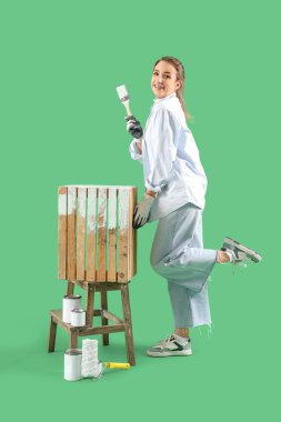 Genç bir kadın yeşil arka planda fırçayla ahşap kutu boyuyor.