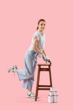 Fırça ve tenekeli genç bir kadın pembe arka planda tabure boyuyor.