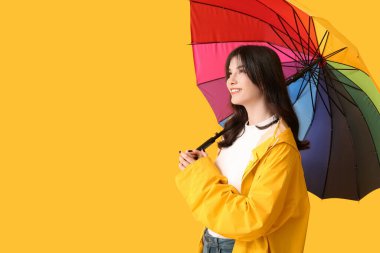 Sarı arka planda açık renkli şemsiyeli yağmurluk giyen genç bir kadın.