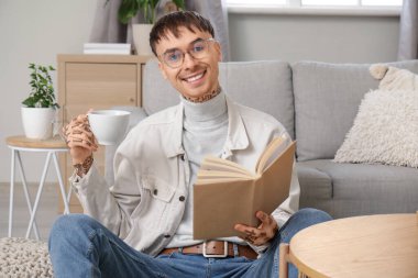 Gözlüklü genç adam elinde çay fincanı ile evde kitap okuyor.