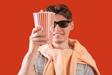 3 boyutlu gözlüklü yakışıklı adam elinde patlamış mısırla kırmızı arka planda kova tutuyor.