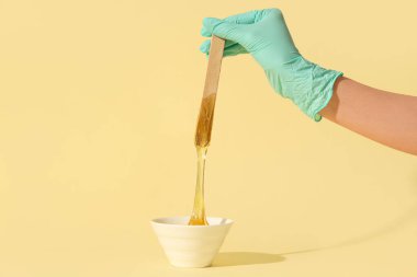 Güzellik uzmanı elinde spatulayla soluk sarı arka planda şekerleme macunu tutuyor.