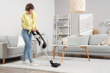 Beautiful young woman cleaning carpet with cordless stick vacuum cleaner in living room
