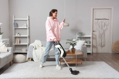 Beautiful young woman cleaning carpet with handheld vacuum cleaner and mobile phone in living room