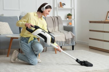 Beautiful young woman in headphones cleaning carpet with handheld vacuum cleaner in living room