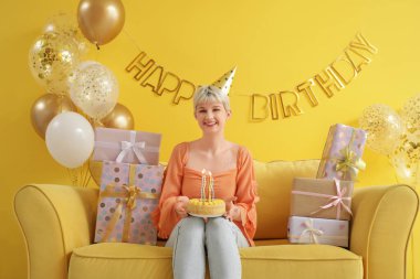 Doğum günü pastası, hediye kutuları ve süslemeleri olan mutlu genç kadın sarı duvarın yanındaki koltukta oturuyor.