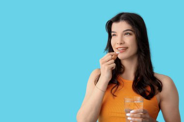 Mavi arka planda bir bardak su ve balık yağı kapsülü olan güzel, genç ve mutlu bir kadın.