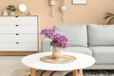 Gri kanepenin yanındaki sehpanın üzerinde bir demet leylak dalı olan oturma odasının içinde.