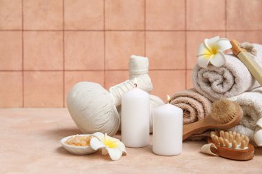 Masaj fırçaları, bitki torbaları, tesisat çiçekleri ve bej masa üzerinde yanan mumlar olan spa malzemeleri.